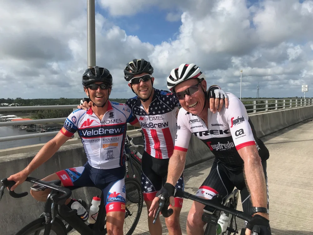 Three smiling cyclists posing together after a group ride on a bridge