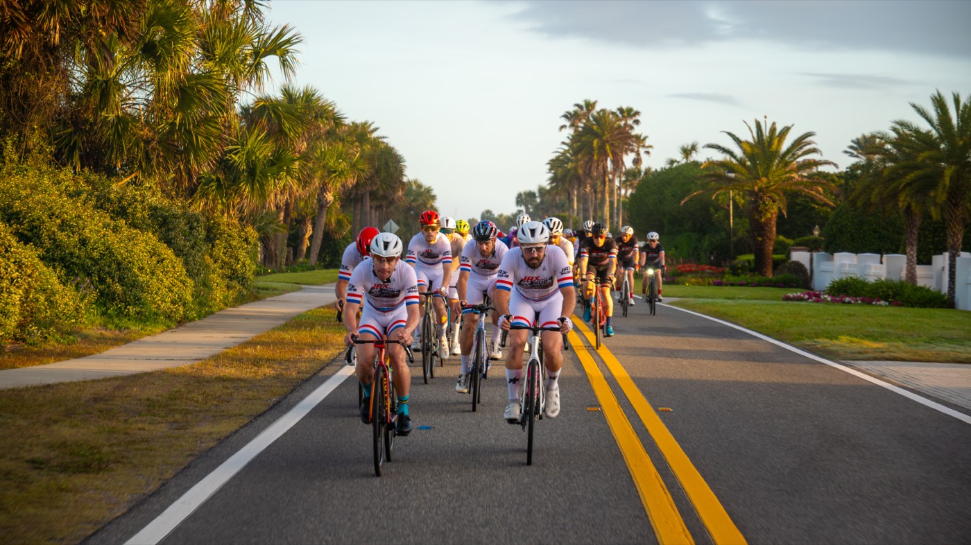 A paceline of riders in Jax Sub 4 kit rolling down a palm-lined road at sunrise.