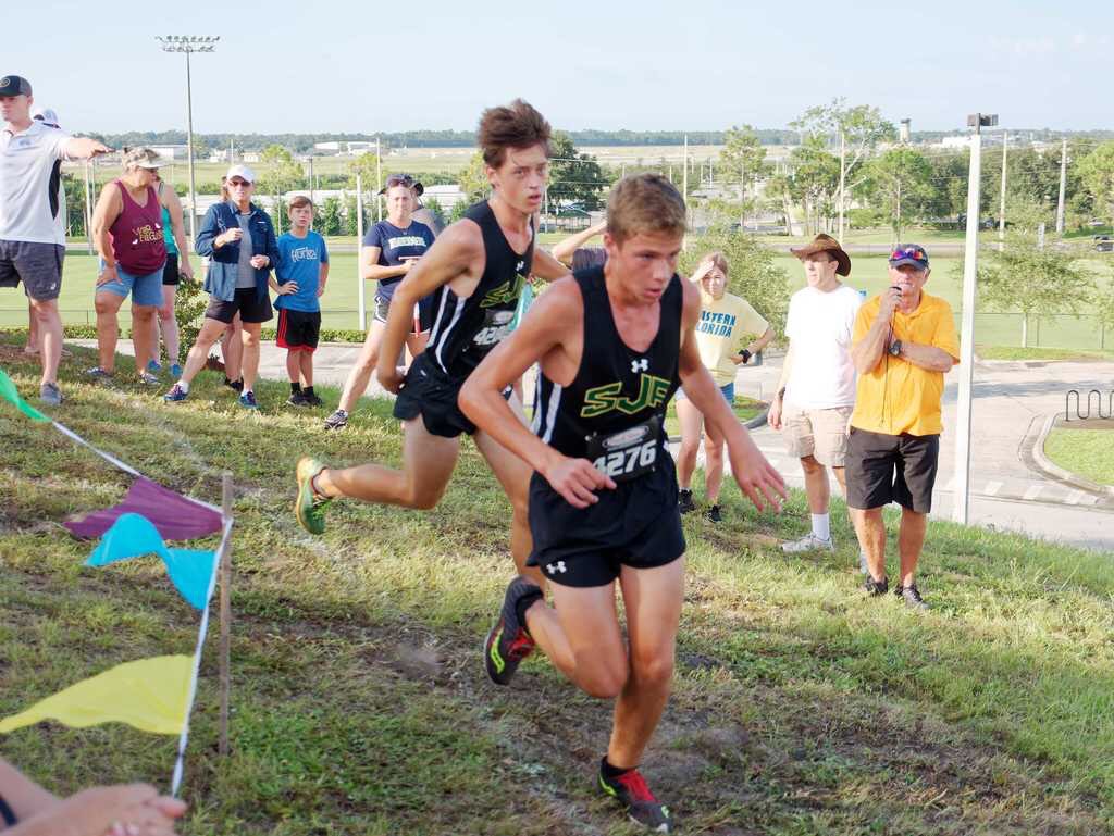 Two SJA cross country runners in green and dark kit charging up a grassy hill during a race as spectators watch from the side.
