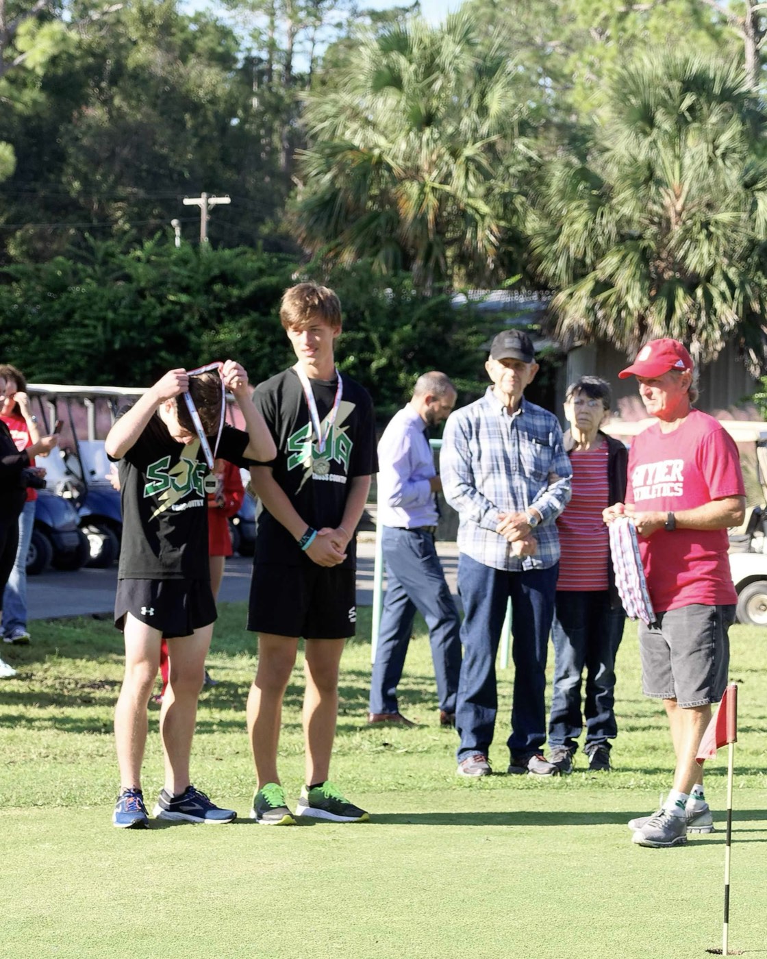 Two SJA cross country athletes at an awards ceremony, one lifting a medal over his head, with palm trees in the background.