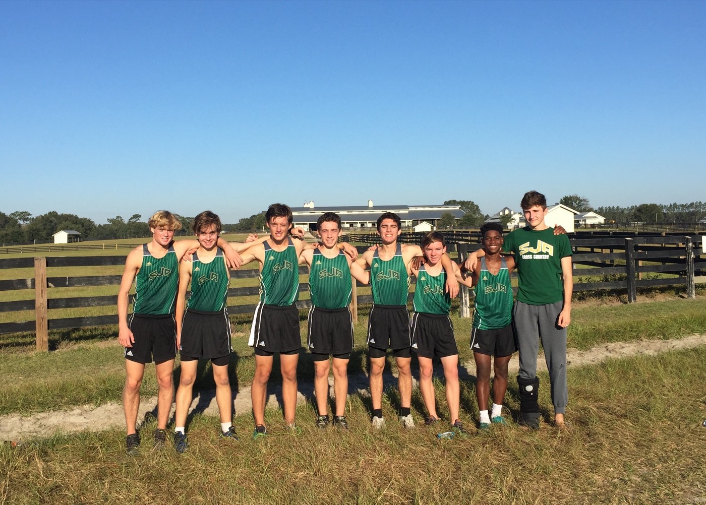 A team of eight boys in green SJA cross country singlets standing arm in arm on a grass field under a blue sky.